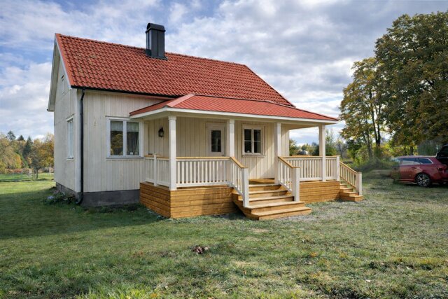 Ett litet gult trähus med rött tegeltak, en veranda med vita räcken och trätrappor, beläget på en gräsmatta med träd i bakgrunden.