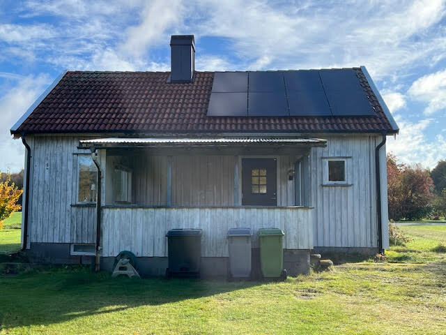 Liten villa med träpanel och tegeltak, veranda framtill, soptunnor nedanför, solpaneler på halva taket, mot en klarblå himmel.
