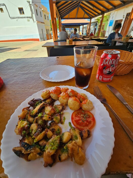 Tallerken med stekt bläckfisk, potatis med sås och tomatskiva, serverad med ett glas Coca-Cola och en burk, på en uteservering.