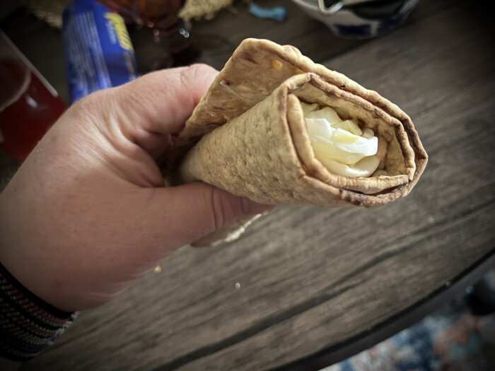 Hand som håller i en macka av tunnbröd med ägg som fyllning, liggandes på ett mörkt träbord.