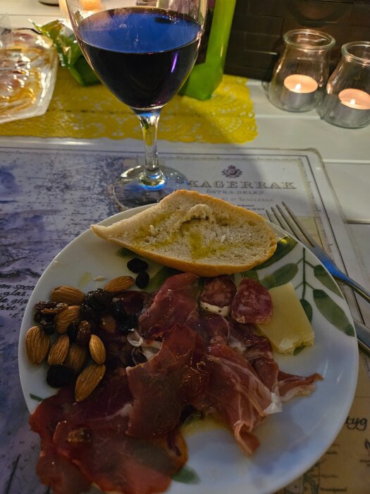 Tallrik med lufttorkat kött, salami, ost, mandlar och russin samt ett glas rött vin och en brödskiva, placerad på ett bord med ljus.