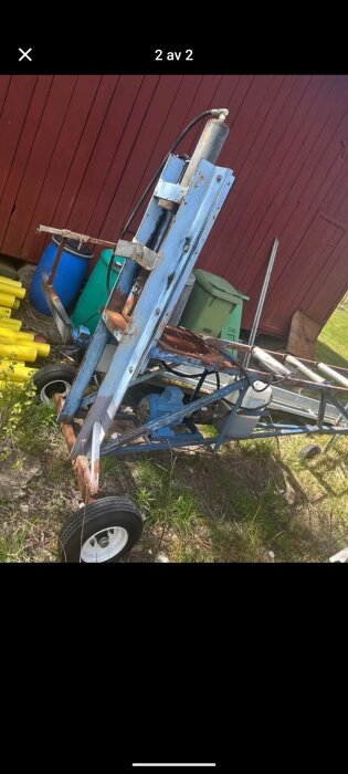 Blå hydraulisk vedklyv på hjul med röda detaljer, omgiven av gröna tunnor och gula rör, står utanför en röd byggnad.
