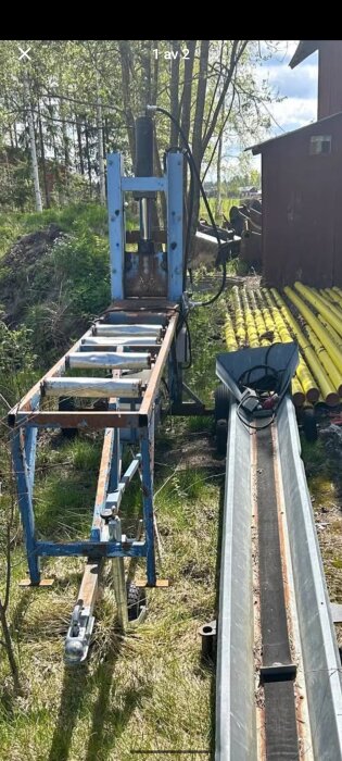 En vedklyv står utomhus bredvid en röd byggnad, omgiven av grönska och gula rörstaplar, med ett transportband i förgrunden.