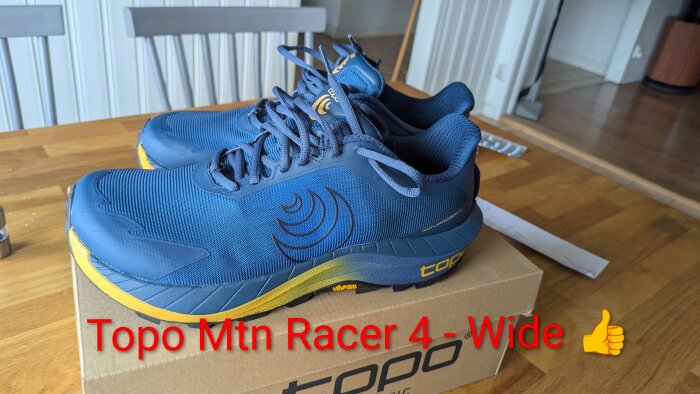 Blåa och gula Topo Mtn Racer 4-skor på en kartong, stående på ett trägolv i ett rum med vitmålade väggar och stolar.