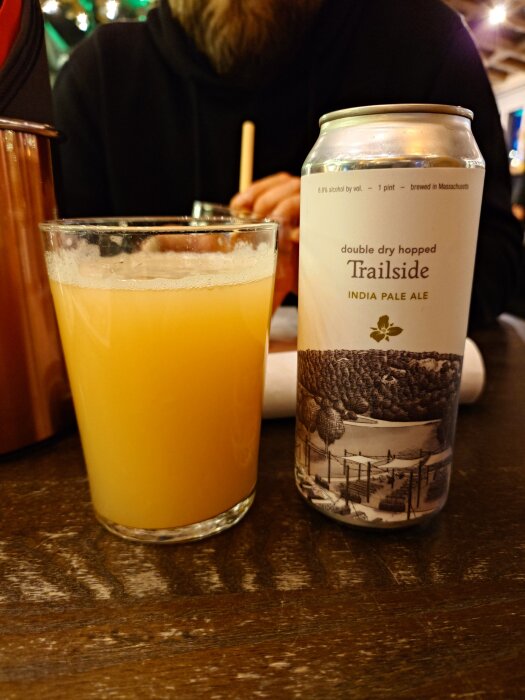 Glas med IPA-bärnstensfärgad öl och Trailside-burk på ett bord, person i bakgrunden.