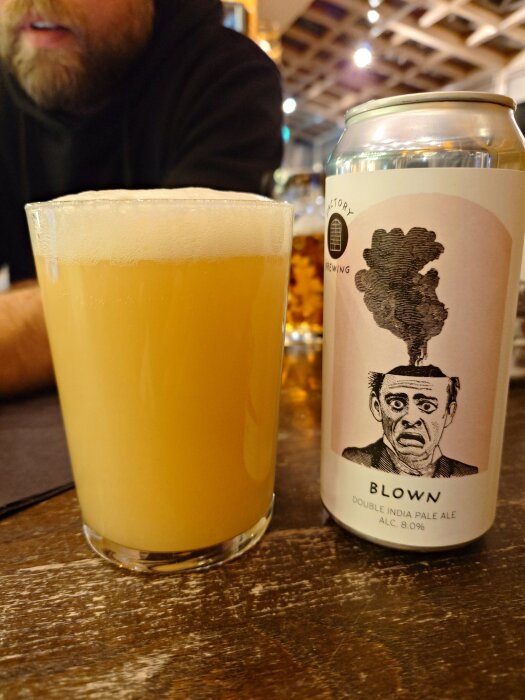 Ett glas ljus öl bredvid en burk märkt "Blown Double India Pale Ale 8.0%" på ett bord i baren.
