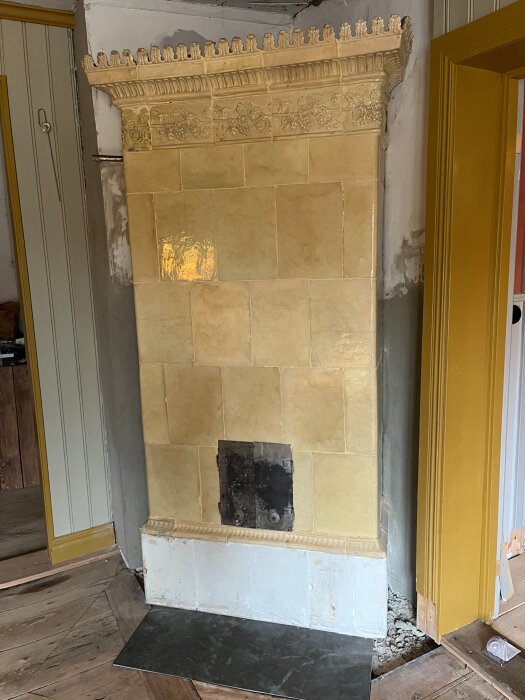 Gammal kakelugn installerad i ett rum, med detaljerat hantverk och gul ton, placerad mot en vägg med omgivande dörrar och träpanel.
