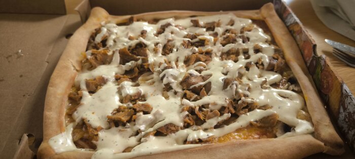 Rektangulär pizza med kebabkött, svamp och vit sås, serverad i en kartong på ett bord.
