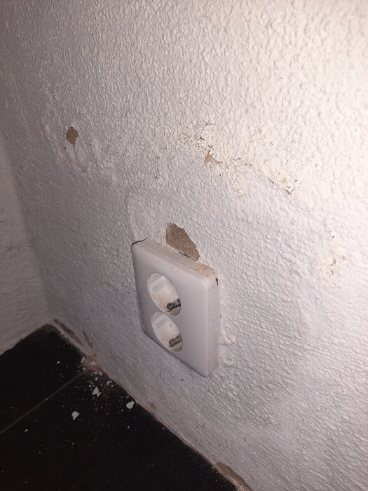 Vägg med avflagad vit färg och skador runt ett vägguttag i en byggnad med tecken på fuktproblem.