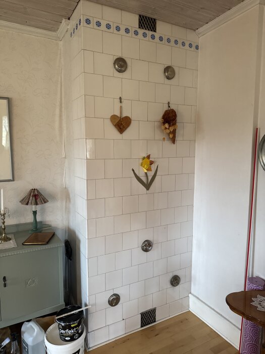 Vit kaklad vägg med blå bård upptill, dekorerad med hjärtformade och blomformade ornament. En hink med färg står på golvet.