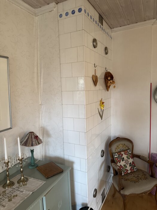 Vit kakelugn i hörn av rum med blommönstrad tapet, dekorationer och en gammal trästol med kudde framför.