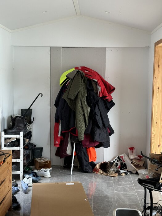 En hall med klädhängare överbelastad med jackor, skor på golvet och en katt liggande i hörnet på en papperspåse.