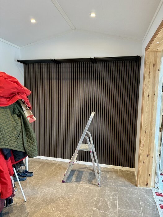 Stege i hall med nyinstallerad mörk ribbpanel på väggen, flera färgglada ytterkläder synliga till vänster.