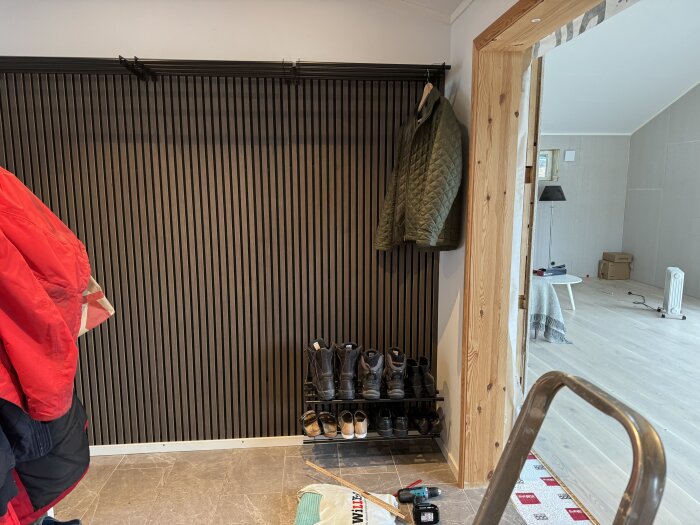 Entré med klädhängare, röda och gröna jackor, skoställ med skor. Nyrenoverat golv, panel och dörrkarm. Verktyg och stege i förgrunden.