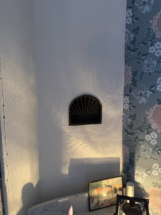 Vägg med vit puts och svart ventil nära murstock, bredvid tapet med blommönster, samt ett bord med en inramad bild och prydnadsföremål.