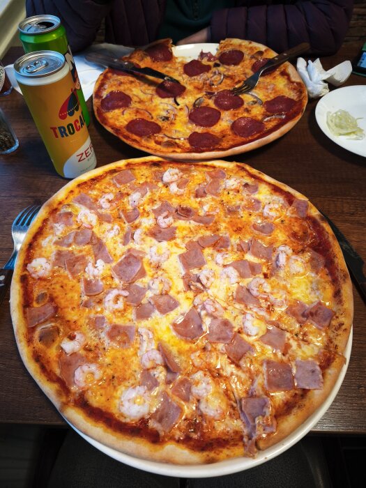 Två pizzor på ett bord med olika toppingar, en med räkor och skinka och den andra med pepperoni. Två burkar läsk på sidan.