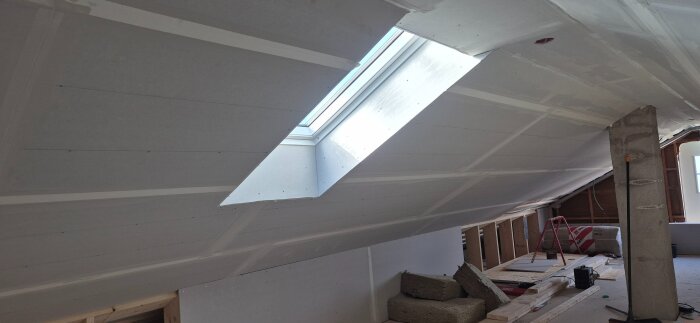 Loft under renovering med installerade gipsskivor, ett takfönster, och byggmaterial utspritt på golvet.