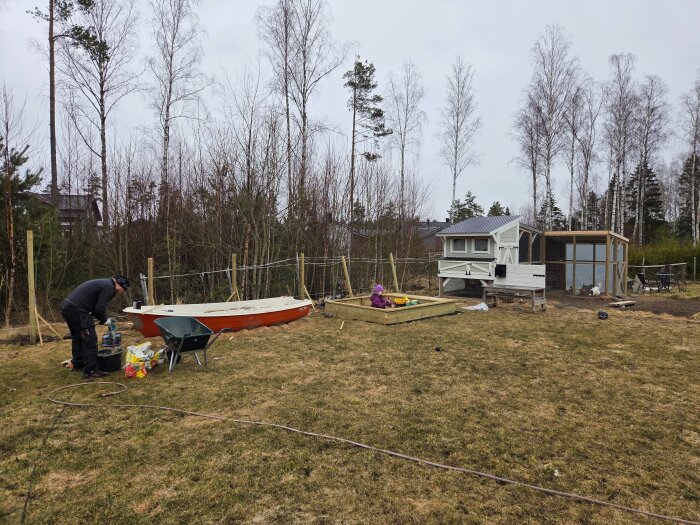 Person arbetar i trädgård med staketstolpar, skottkärra vid en liten båt; lekande barn i sandlåda, hönshus och inhägnad i bakgrunden.