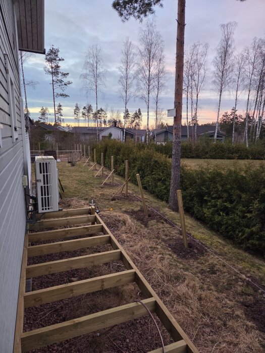 Nybyggda staketstolpar och grund för trädäck vid husets sida, med skog och hus i bakgrunden i solnedgångens sken.