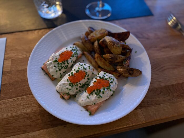 Tallrik med två bitar lax toppade med gräddsås, rom och gräslök, serverade med rostad potatis på ett mörkt träbord.