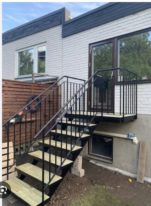 Svart trappa med metallräcke som leder upp till en altandörr på ett hus av vit tegel.