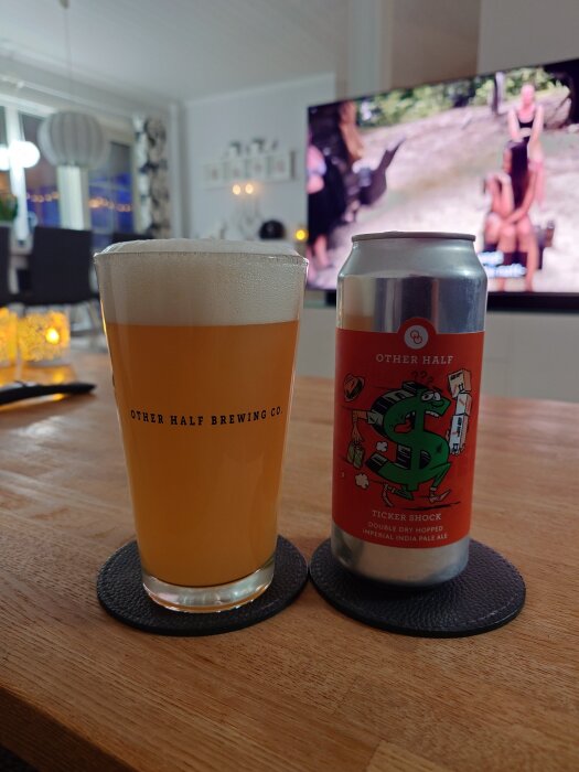 Ett glas öl och en ölburk med etiketten "Other Half Brewing Co." står på ett underlägg på ett träbord, med en TV i bakgrunden.