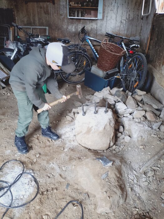 En person i keps och arbetskläder använder en slägga för att dela sten i en ladugård fylld med cyklar och verktyg.