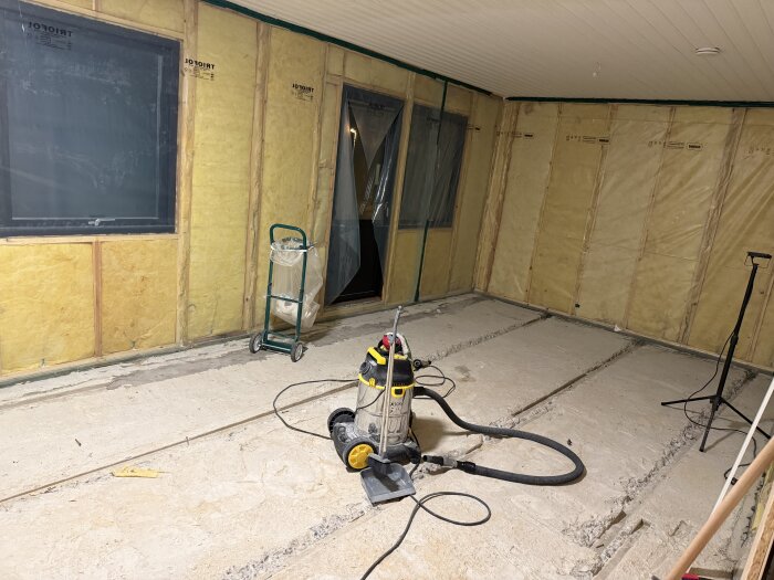 Rum under renovering med exponerade isolerade väggar och en betongslipmaskin på golvet. Verktyg och plats för nya golvytor syns också.