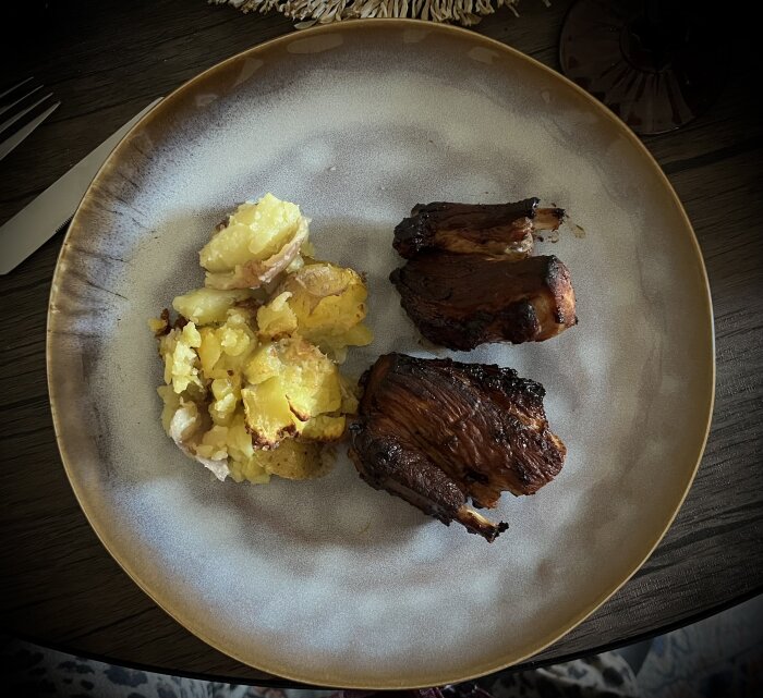 Tallrik med glazeade revbensspjäll och smashed potatoes med vitlökssmör på ett mörkt bord.