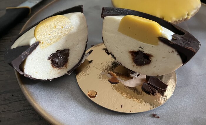 Påskbakelse delad i två halvor med chokladöverdrag och fyllning av vit mousse, mörk kräm och gul topp på en tallrik.