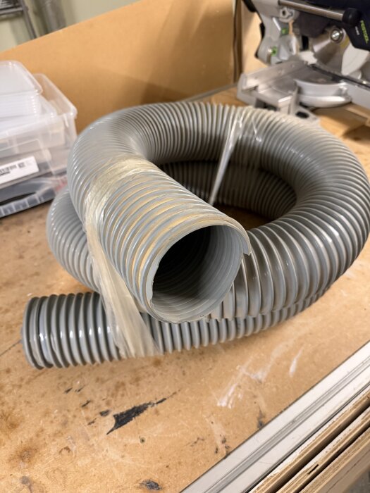 Grå flexibel plastslang 3 m lång med innerdiameter 102 mm, placerad på en arbetsbänk omgiven av verktyg och förpackningar.