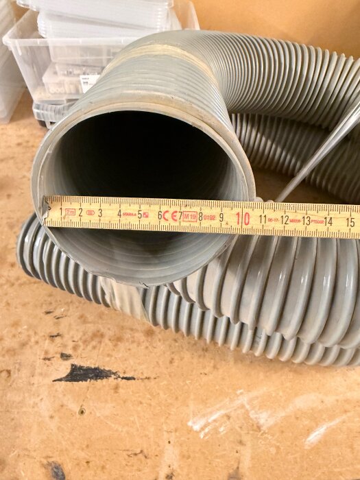 Grå flexibel slang med en linjal ovanpå som visar dess diameter på cirka 11 centimeter, placerad på ett arbetsbord.