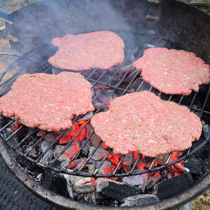 Grillning av fyra biffar av nötfärs med kryddor på kolgrill, glödande kol syns under gallret.