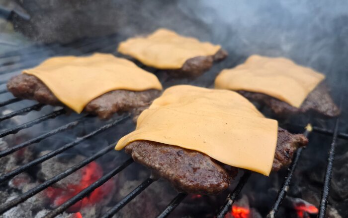Hamburgare av nötfärs med ostskivor på en grill, omgärdade av rök och glödande kol.