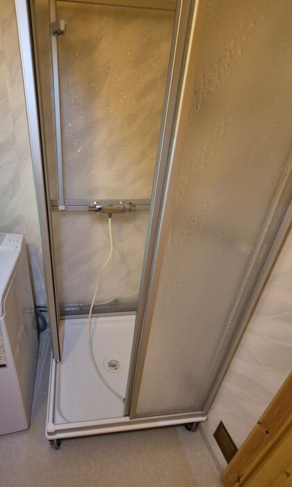 Duschkabin med plastpaneler och metallram i ett badrum med plastmatta och synliga rör, bredvid en vit tvättmaskin.