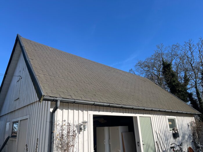 En vitmålad garagebyggnad med sadeltak täckt av shingel ses från sidan, med blå himmel och träd i bakgrunden.