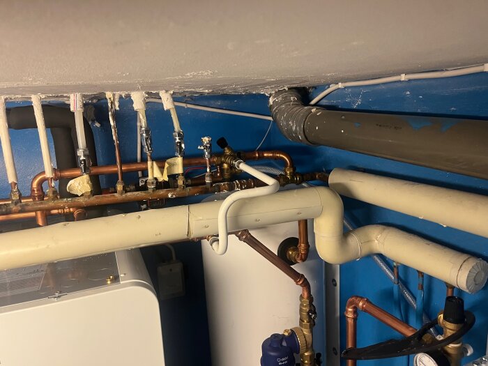 Rör och ventiler i ett värmesystem med koppar- och pvc-rör i en teknikrumsmiljö, blå vägg i bakgrunden.