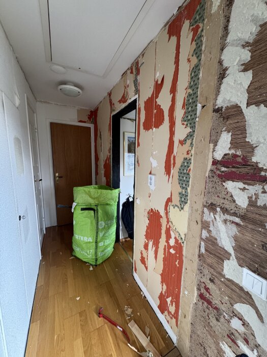 Hallen under renovering med flera lager gammal tapet framträdande, verktyg på golvet och en grön avfallssäck synlig.