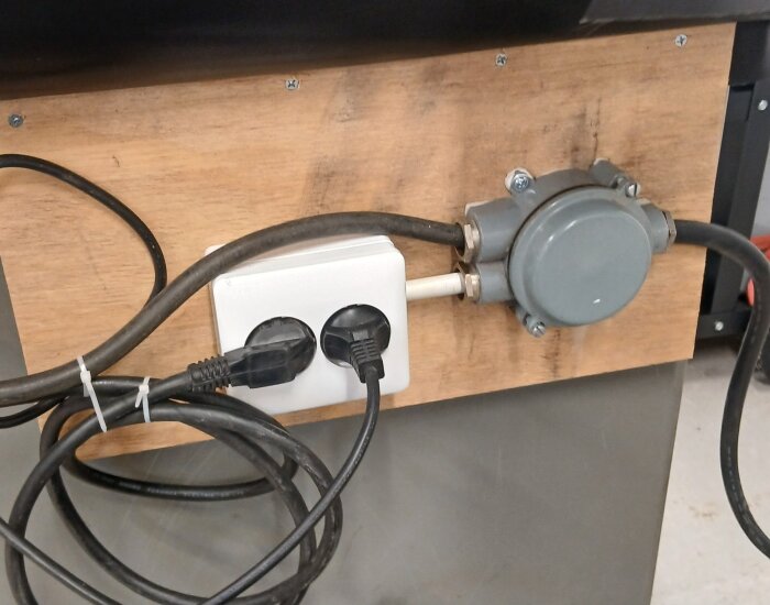 Eluttag monterat på träskiva med flera elkablar anslutna, som en del av arbetsbänkens installation i ett garage.