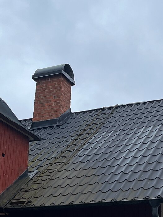Röd tegelstensskorsten på svart plåttak med stege men utan arbetsplattform, mot en molnig himmel.