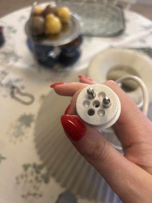 Gammal lamppropp i vit plast med fyra hål och två stift hålls mellan fingrarna, mot en bakgrund av ett rum och en fruktskål.