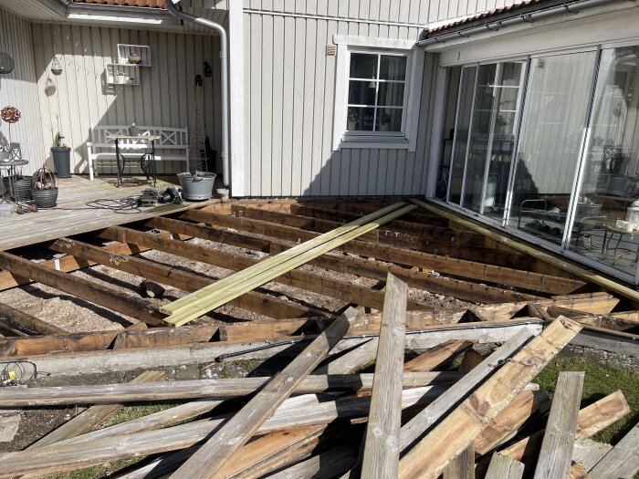 Trädäck under renovering med brädor, reglar och verktyg. Husets fasad och fönster syns i bakgrunden.