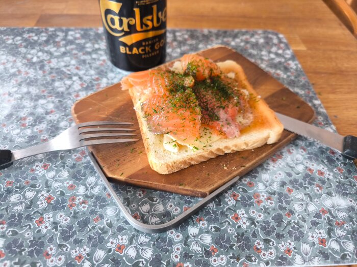 En toast med hemgravad lax och Västerbottensost på en träbricka, med en gaffel och kniv bredvid samt en Carlsberg Black Gold öl i bakgrunden.