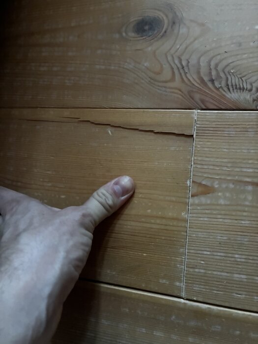Sprucken golvplanka i trä med längsgående spricka vid en skarv. En hand visas som pekar på skadan i samband med renoveringsdiskussion.