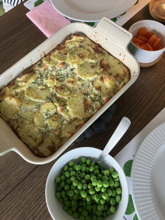 En laxpudding i ugnsform och en skål med gröna ärtor på ett dukat bord med tallrik och ljus.