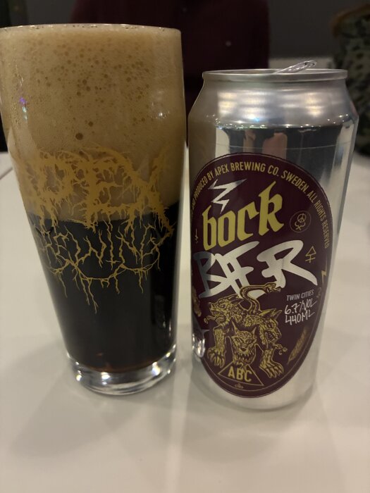 Ett glas med mörk öl och skummig topp bredvid en burk med etikett från Apex Brewing Co. och texten "bock beer".
