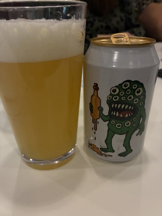 Ölglas med skummande bärnstensfärgad öl och en ölburk med tecknad flerögd figur på ett bord.