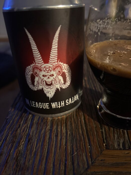Ölburk med etiketten "In league with satan" från Snausvarve gårdsbryggeri, bredvid ett glas stout på ett träbord.