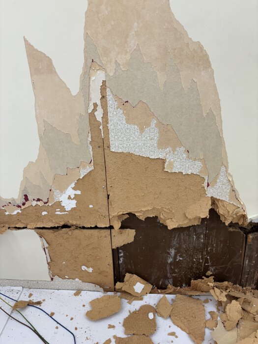 Gammal tapet delvis borttagen från vägg, med bitar som ligger på golvet, exponerar underliggande yta i rummet som förbereds för renovering.