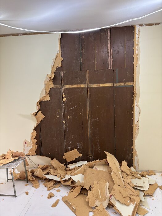 Riven tapet avslöjar träpanel bakom väggen i ett renoveringsprojekt. Fragment av gammal tapet ligger spridda på golvet.